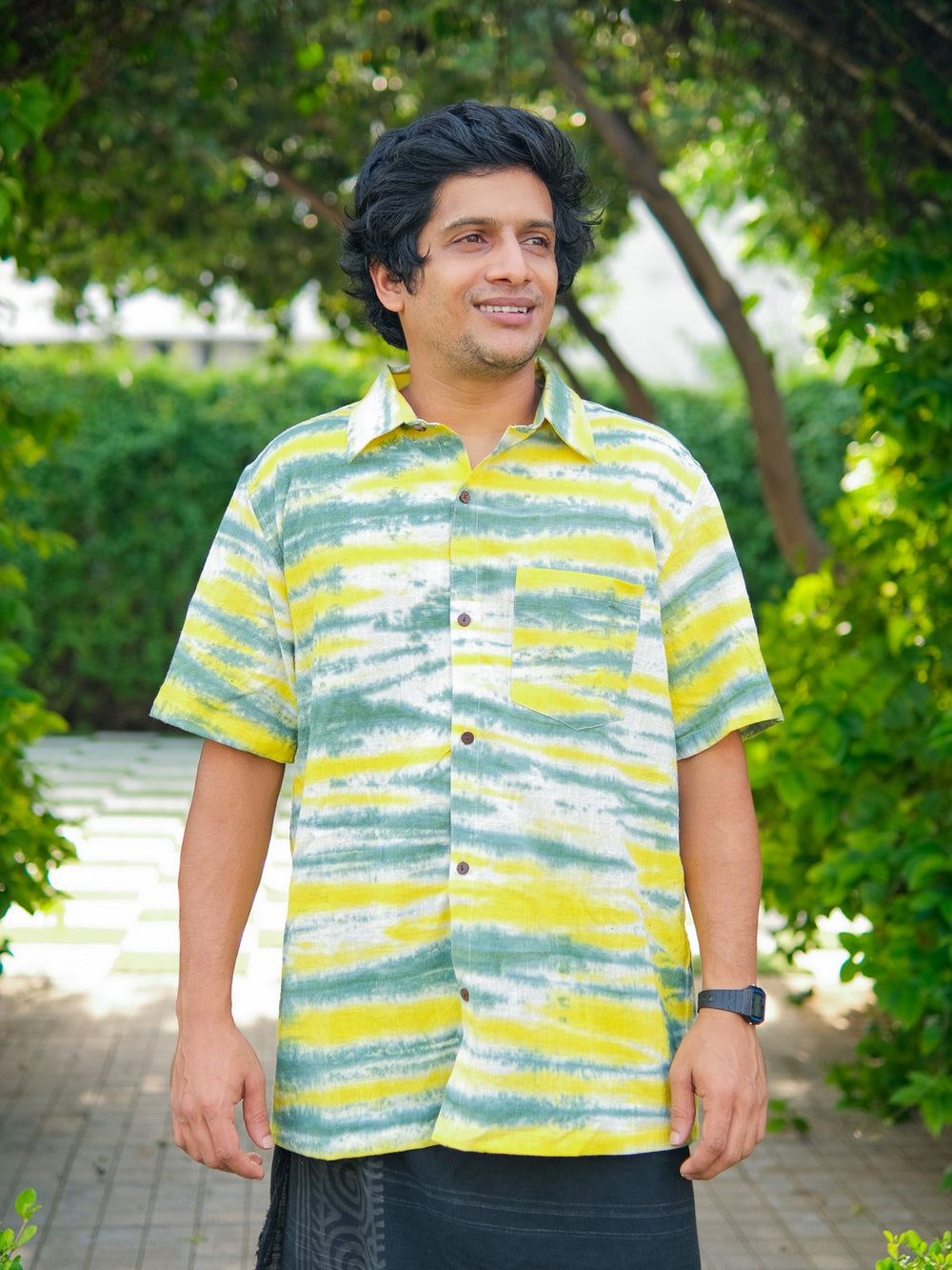 A man stands outdoors, smiling, wearing the Tusha Shirt—a yellow, green, and white striped top made from premium quality cotton. Green trees and sunlight create a natural, vibrant background.