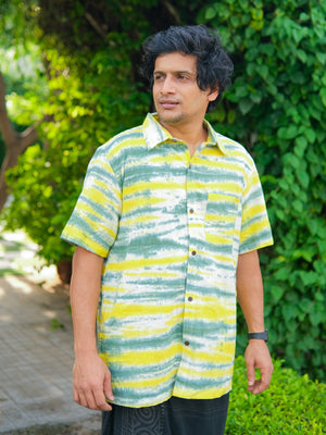 A man stands outdoors on a pathway surrounded by greenery and trees, wearing the Tusha Shirt—a premium quality cotton shirt featuring green, yellow, and white stripes.
