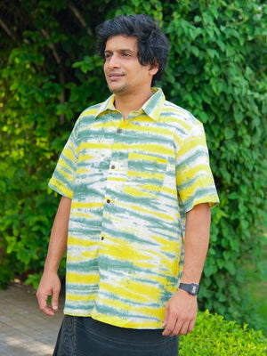 A man with dark hair, dressed in a yellow and green striped Tusha Shirt crafted from premium quality cotton, stands outdoors before lush greenery, wearing a subtle smile.