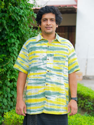 Standing outdoors, a person wears the Tusha Shirt—crafted from premium quality cotton with yellow, green, and white stripes—paired with a black watch and dark pants. Lush green bushes and a building can be seen in the background.