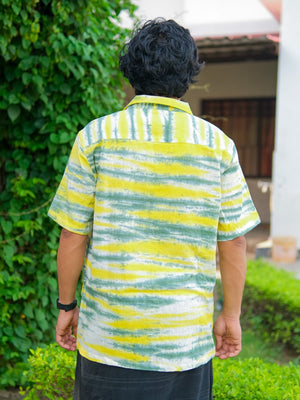 A person with short black hair is seen from behind, standing outdoors near greenery and a building, wearing the Tusha Shirt in a yellow and green tie-dye pattern made from premium quality cotton, paired with a black watch.