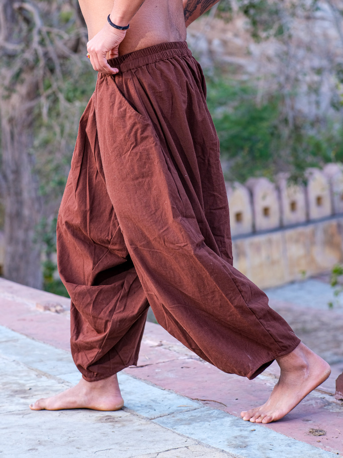 A barefoot person walks outdoors wearing Sirwal pants with a modern twist. The scene includes a stone pathway, lush greenery, and a weathered stone wall in the background.