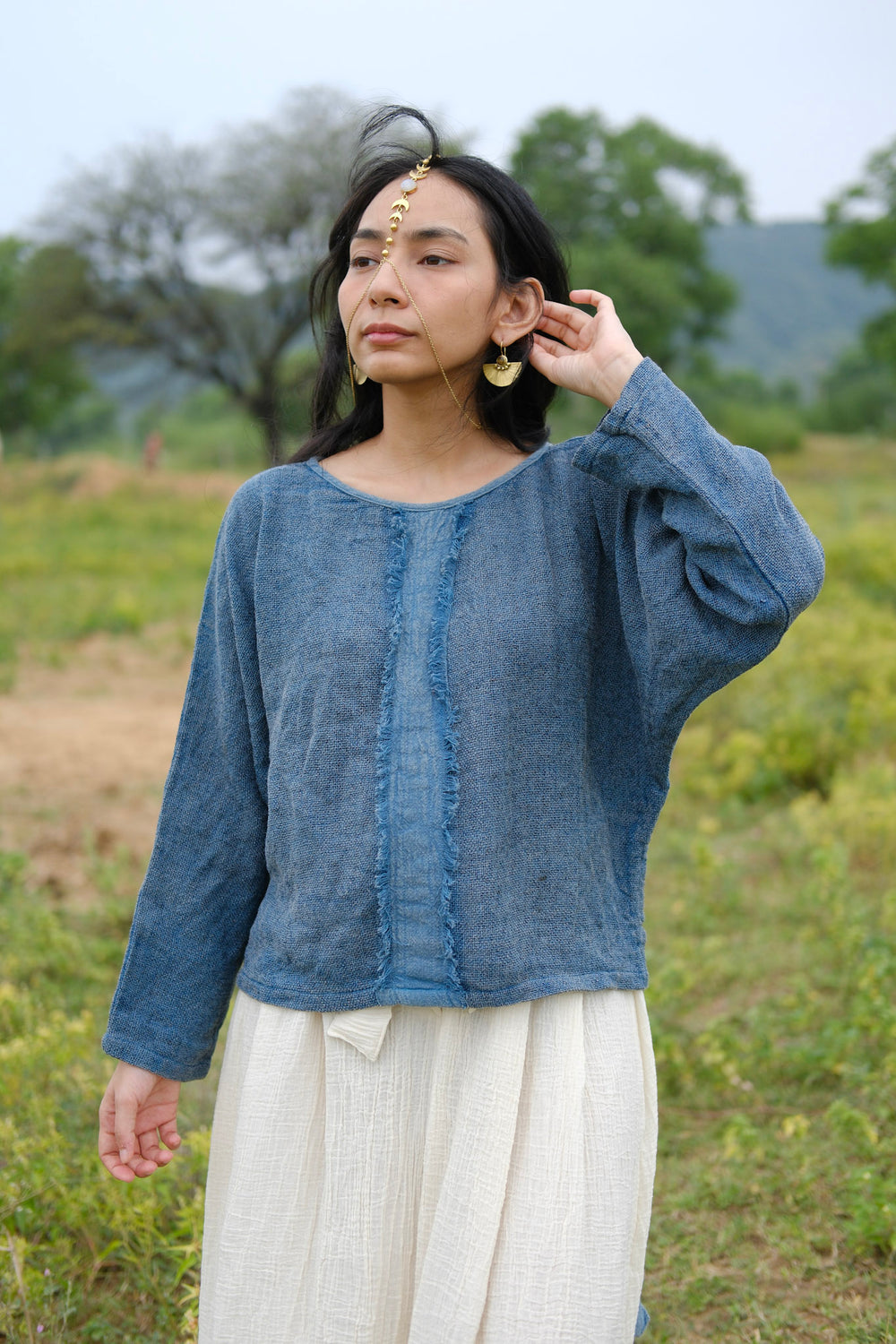 A woman stands outdoors, framed by green trees and hills, wearing the Bohemian Long-Sleeve Top in blue paired with a white skirt. She accessorizes with gold earrings and a forehead ornament, gently touching her hair and looking away—a perfect scene for effortless resort wear.