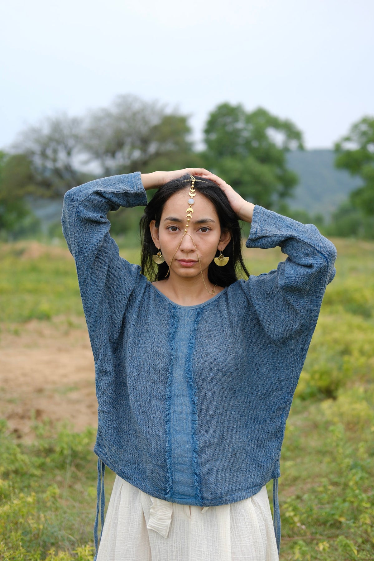 Wearing the Bohemian Long-Sleeve Top paired with a light skirt, a woman stands outdoors with her hands on her head, adorned in gold jewelry and a delicate forehead pendant. The lush greenery and rolling hills behind her create a serene setting, highlighting the top as an ideal choice for resort wear.