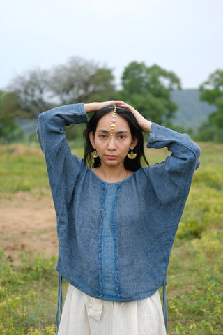 Wearing the Bohemian Long-Sleeve Top paired with a light skirt, a woman stands outdoors with her hands on her head, adorned in gold jewelry and a delicate forehead pendant. The lush greenery and rolling hills behind her create a serene setting, highlighting the top as an ideal choice for resort wear.
