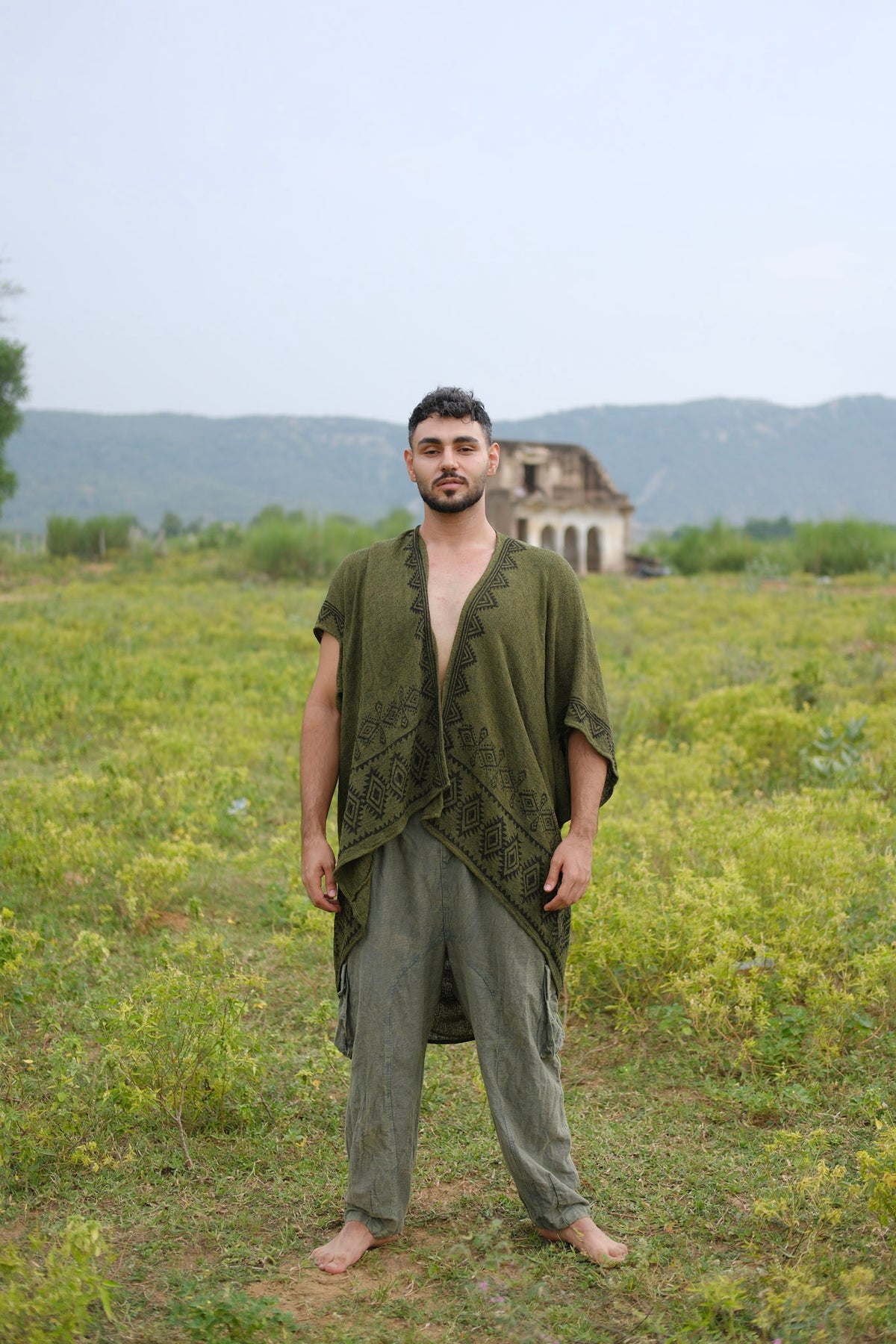 Barefoot on the grass, a person wears loose olive-green clothing featuring a geometric pattern and completes the look with the stylish Miraya Shrug. Greenery surrounds them, set against an old, weathered building and distant mountains beneath a pale sky.