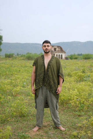 Barefoot on the grass, a person wears loose olive-green clothing featuring a geometric pattern and completes the look with the stylish Miraya Shrug. Greenery surrounds them, set against an old, weathered building and distant mountains beneath a pale sky.