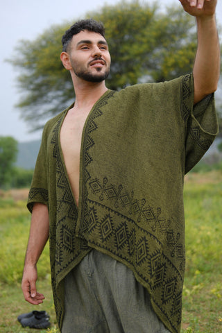 A man wearing the Miraya Shrug, an olive green patterned poncho reminiscent of a stylish shrug, stands outdoors in a grassy area, smiling and gesturing with his right hand. A tree and hills can be seen in the background.