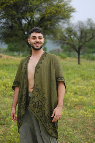 A man stands smiling in a grassy field, dressed in an open, patterned green Miraya Shrug paired with loose pants. Trees and a cloudy sky form the backdrop, enhancing the relaxed yet stylish vibe of his Miraya Shrug ensemble.