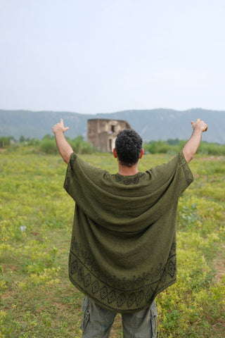 Wearing a Miraya Shrug with a green pattern, someone stands in a grassy field, arms raised and pointed toward a distant stone building and rolling hills beneath a cloudy sky.