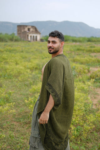 A man wearing a green robe and the Miraya Shrug stands in a grassy field, looking back over his shoulder, with mountains and an old stone building in the background beneath a cloudy sky.