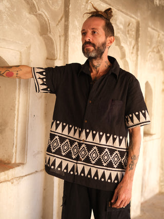 A bearded man with a topknot stands against a textured wall, wearing the Chakor Shirt with black fabric featuring geometric white block-print-like patterns. Tattoos decorate his arms and neck as he gazes thoughtfully into the distance.