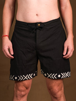 Wearing Tribal Shorts in black with white geometric hem patterns, made from comfortable cotton, the person stands against a brown background with relaxed arms and only their torso and legs visible.