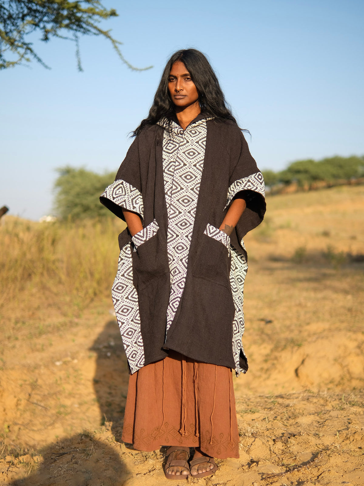 A person wears a black Neer Poncho with white diamond-patterned trim and front pockets over a long brown skirt and sandals.