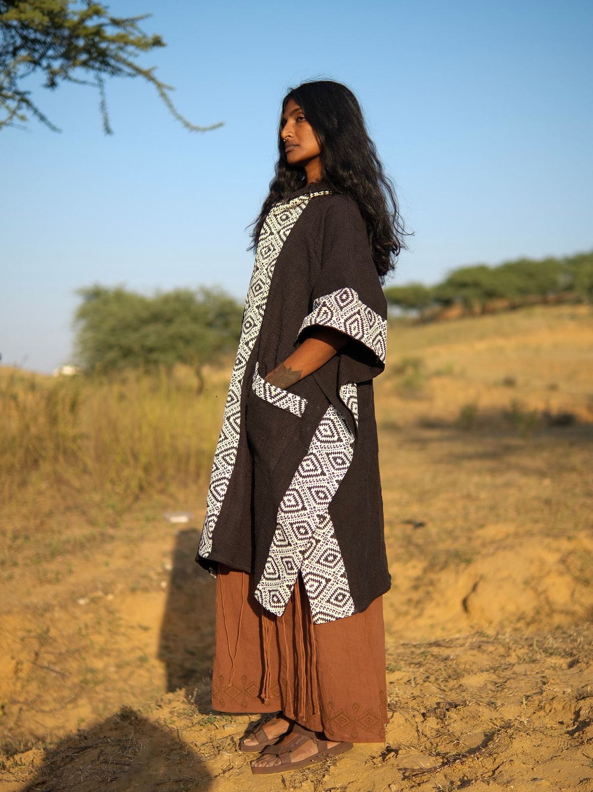 A person wearing a black Neer Poncho with white geometric patterned trim over a brown skirt standing in a desert landscape.