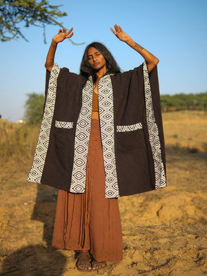 A person wearing a black open-front Neer poncho with a white geometric patterned border and matching pocket details, standing in an outdoor setting with arms raised.