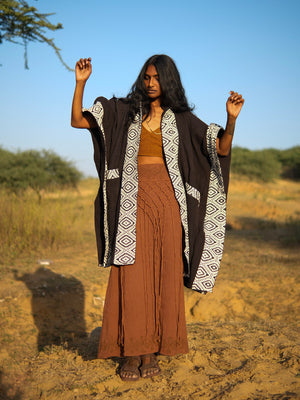 A person wears an open black Neer poncho with a white geometric pattern along the edges over a brown crop top and a long, fringed brown skirt.