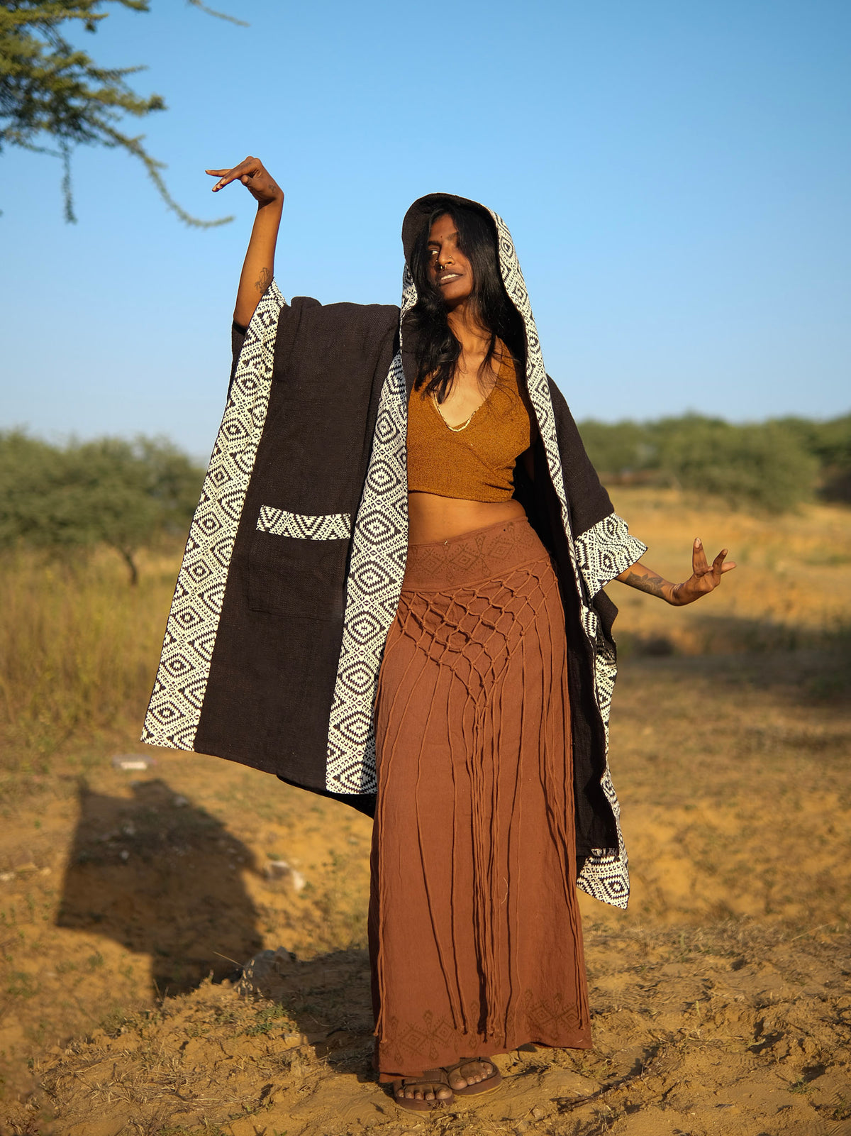A person in a black Neer Poncho with a white geometric trim, worn over a brown crop top and a long fringe skirt, standing in a desert landscape.