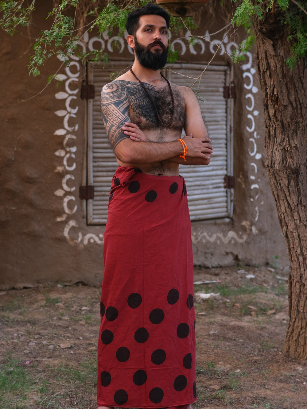 A bearded, tattooed man stands outdoors with his arms crossed, wearing a Prabhav Dhoti distinguished by bold black polka dots on red—showcasing sustainable design and exquisite hand block print craftsmanship—in front of a brown wall adorned with white motifs and a closed shutter, next to a tree.