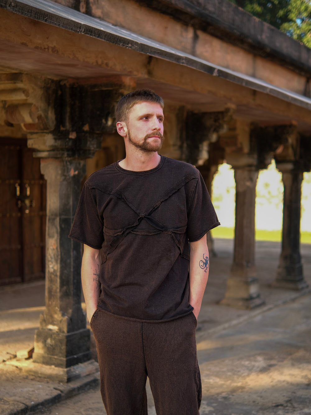 A man with short hair and a beard stands outdoors in front of ancient stone pillars, wearing the Noah T-Shirt and dark pants, with his hands in his pockets, looking to the side.