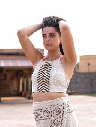 A person with long dark hair, wearing a white Mathu Crop Top and a matching Rajasthani hand block printed skirt with geometric patterns, pulls their hands behind their head. They stand outdoors under a clear sky with structures visible in the background.