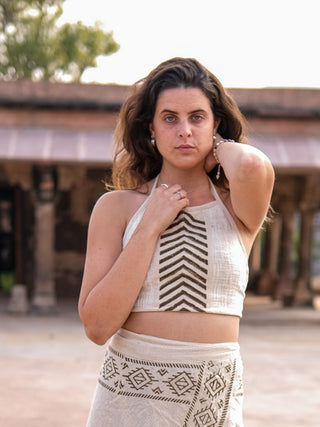 A woman showcases her boho flair in a Mathu Crop Top and matching skirt with geometric Rajasthani hand block prints. With long brown hair cascading naturally, she poses with one hand on her neck in front of a building featuring classic columns.