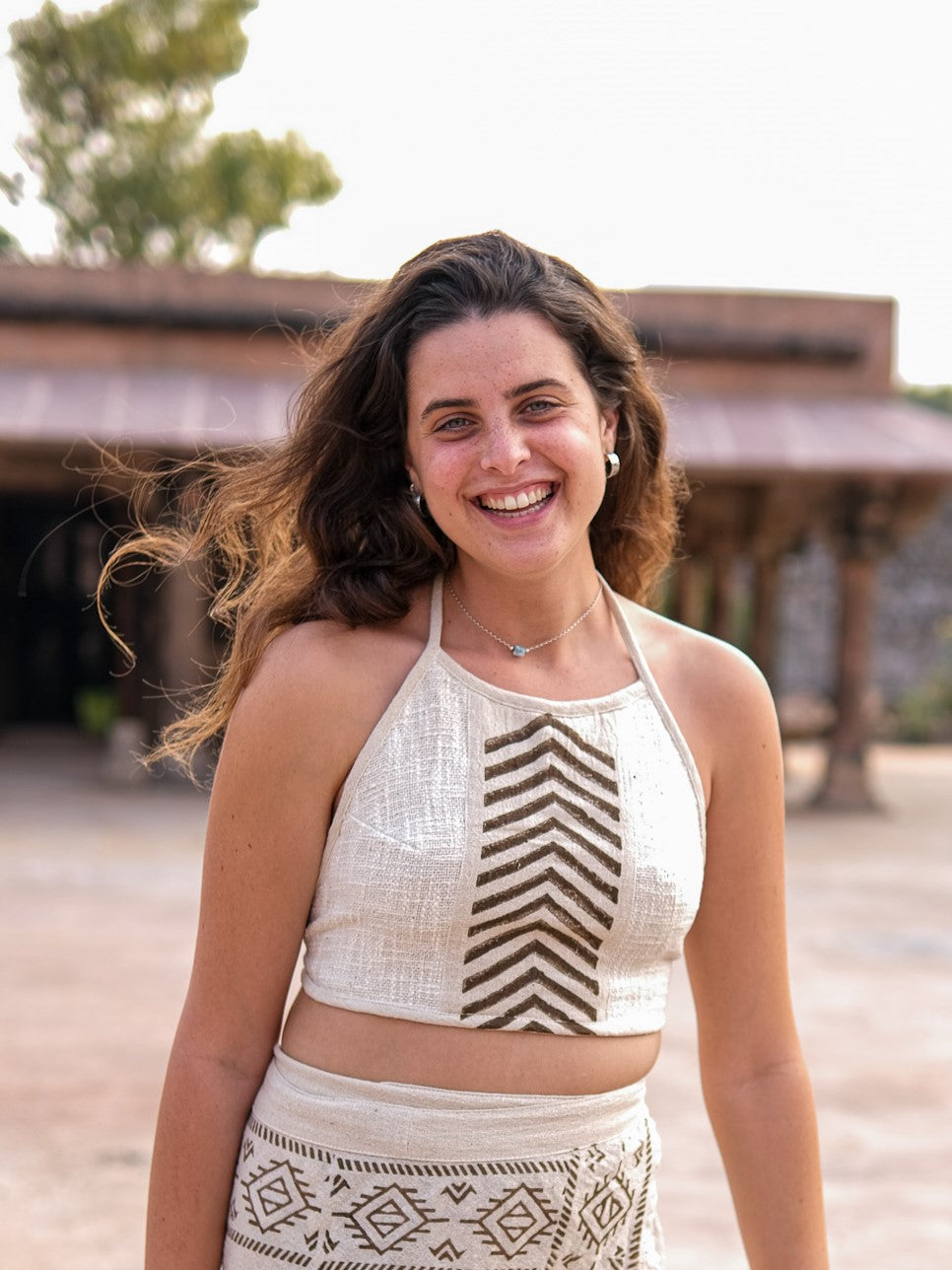 A woman stands outdoors, showcasing the Mathu Crop Top in a boho style paired with a patterned skirt. Her long, wavy hair flows gracefully against a rustic backdrop of trees and an old building.