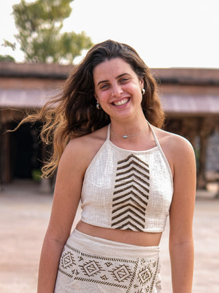 A woman exudes effortless boho charm outdoors, smiling in a Mathu Crop Top with a matching skirt. Her long, wavy hair cascades gracefully against a rustic building and lush trees background.