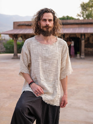 A man with long curly hair and a beard stands outdoors, wearing a Baggy Knit T-shirt in boho style with dark pants. The background shows a rustic building with arches and trees under a clear sky.