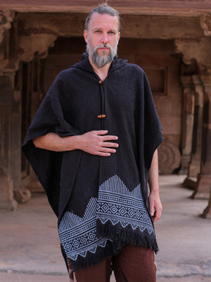 A bearded man stands outdoors, modeling the Swing Shawl—a black poncho featuring white geometric patterns and fringe along the bottom—paired with brown pants. He rests one hand on his chest and looks at the camera.