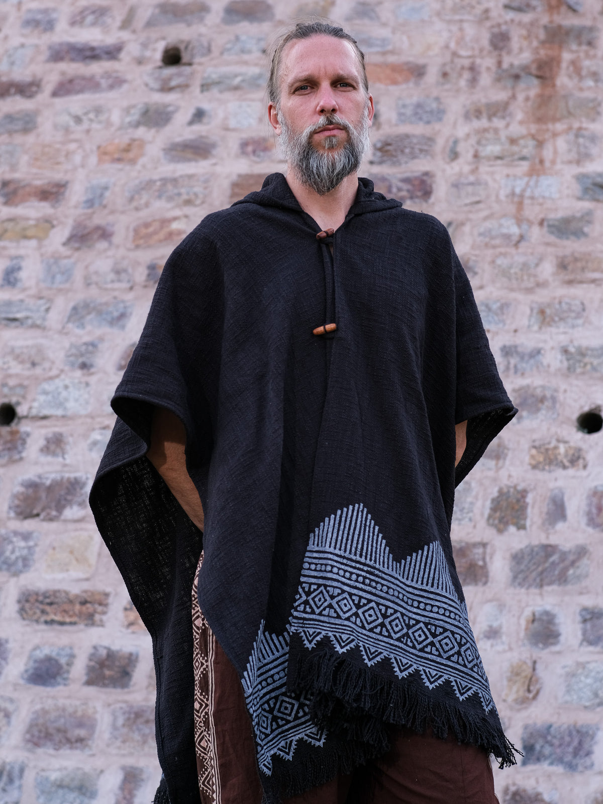 A bearded man stands against a stone wall, wearing a black hooded poncho adorned with light blue geometric patterns along the hem. Draped over his shoulders is the black Swing Shawl, complementing his outfit perfectly. With his hands tucked into his pockets, he gazes calmly at the camera.
