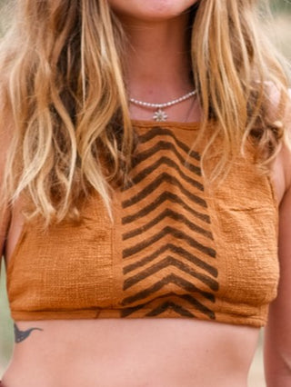 A woman with long blonde hair wears the Mathu Crop Top, a boho-style burnt orange halter with a black chevron center. She sports a star-shaped necklace and has a small tattoo on her left ribcage. Her face is not visible in the image.