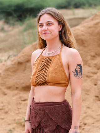 A person with long hair stands smiling outdoors, wearing the Mathu Crop Top paired with a brown textured skirt. The upper arm shows a portrait tattoo. Sandy terrain and green plants create a boho style vibe.