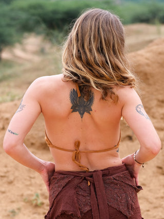 A person with shoulder-length hair stands outdoors in a Mathu Crop Top, showcasing a detailed upper back tattoo and additional arm tattoos, all set against an earthy, boho-style landscape.