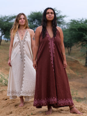 Two women stand barefoot outdoors on sandy ground, surrounded by green trees and a cloudy sky. They wear Hanvi maxi dresses—sleeveless styles featuring geometric patterns, crafted from sustainable cotton that blends eco-friendly fashion with elegant design.
