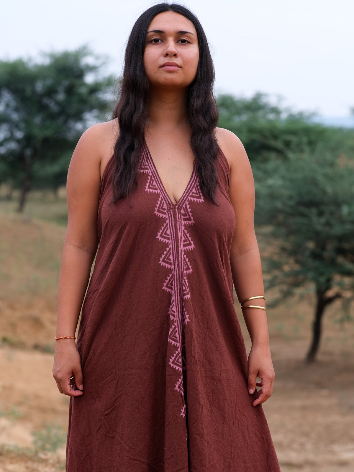 A person with long dark hair stands outdoors, wearing the Hanvi maxi dress—a sleeveless, deep v-neck brown dress featuring pink block print geometric patterns—paired with gold bangles and a red thread bracelet, set against a backdrop of trees and a grassy field.
