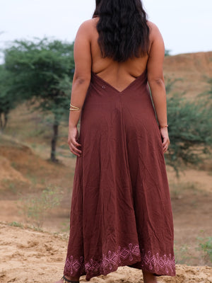 A woman with long dark hair stands outdoors, her back to the camera, wearing the Hanvi maxi dress in brown—a backless style featuring a decorative hem and crafted from sustainable cotton. Desert terrain and green trees form the background.