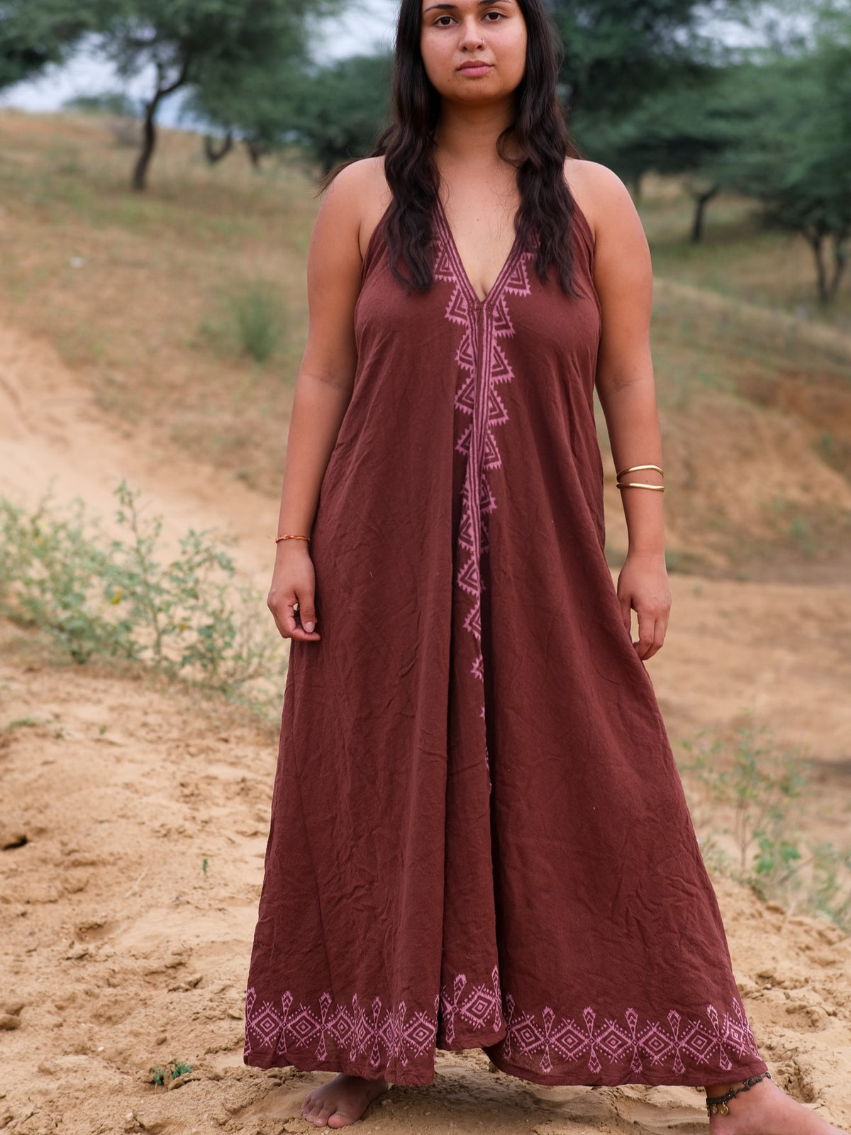 A woman stands barefoot on sandy ground outdoors, wearing the brown Hanvi maxi dress—crafted from sustainable cotton and adorned with pink geometric embroidery inspired by block print patterns—with trees and grassy hills in the background.