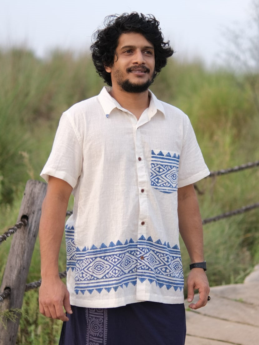 A man with curly hair and a beard is standing outdoors amidst greenery, wearing the Bohemian Tribal Shirt—a short-sleeved, cream cotton shirt adorned with blue geometric patterns.