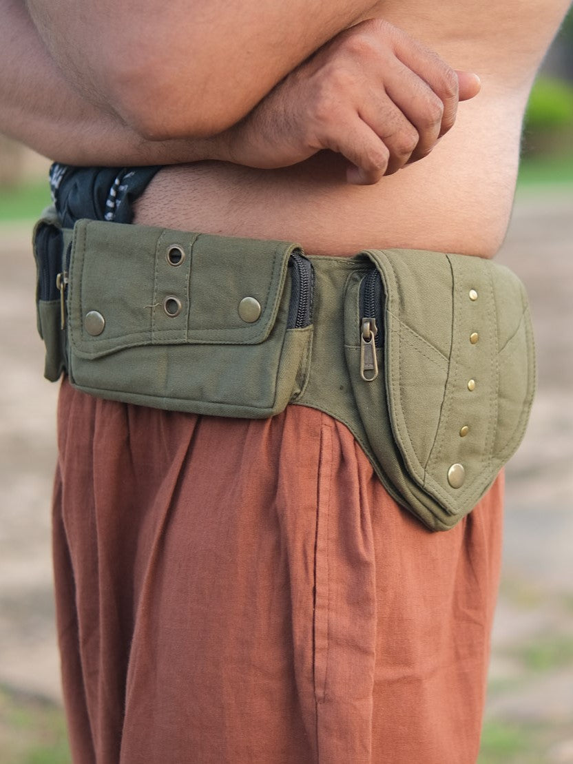 A person wearing brown pants stands outdoors with arms crossed over their chest, showcasing the Verdant Multi-Pocket Utility Belt Bag, which features multiple zippered and buttoned pockets for practical travel use.