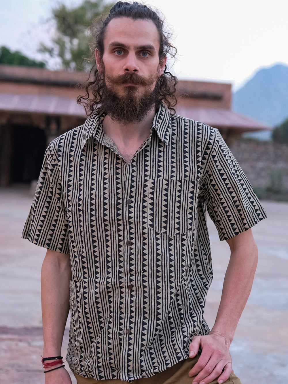 A man with a beard and long curly hair stands outdoors, wearing the Tribal Triangle shirt in black. Blurred buildings and trees can be seen in the background.