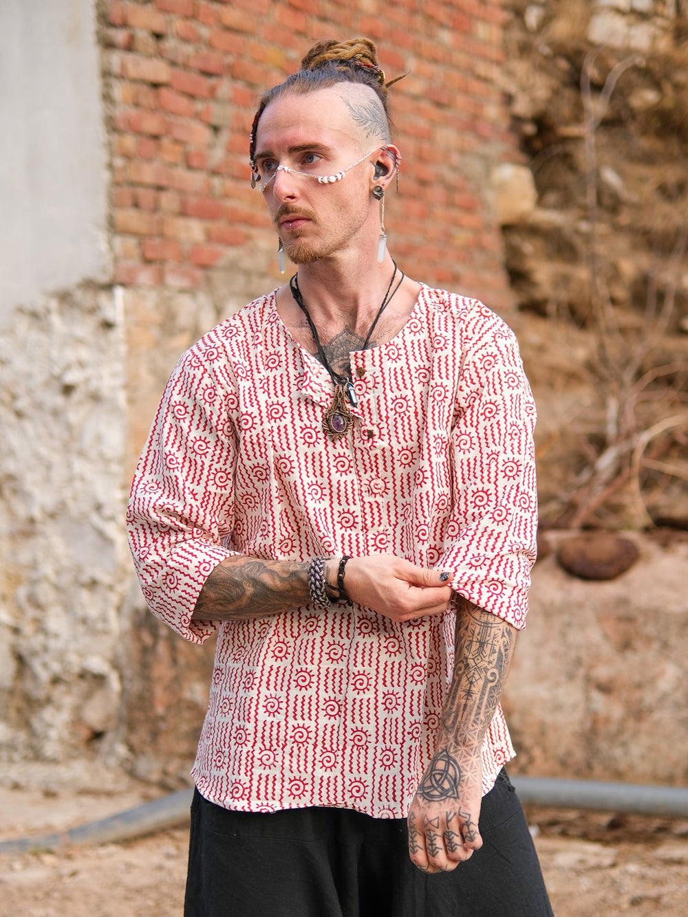 A tattooed man with facial piercings and dreadlocks stands outdoors against a rustic wall, wearing a 100% cotton, hand block printed Moche Kurta, black pants, necklaces, light face paint, and his hair tied up.