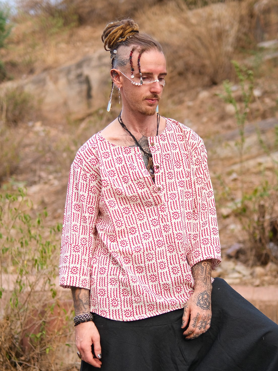 A person with dreadlocks, facial jewelry, and tattoos wears a hand block printed red and white Moche Kurta made from 100% cotton with black pants, standing outdoors in a dry, grassy area with rocks behind them.