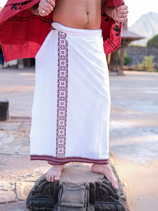 Barefoot on a stone platform, a person exudes elegance in a stylish Chakrot Dhoti with red embroidered borders. Partially covered by an open red shirt, the dhotis high-quality fabric enhances the serene outdoor setting of blurred structures and trees.