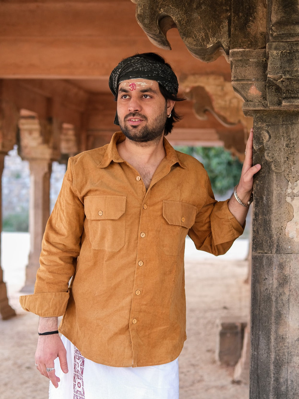 A man in a stylish mustard Fane Shirt, paired with a white traditional garment and black headscarf, stands beside a carved stone pillar in a rustic outdoor setting, marked by a red tilak on his forehead.