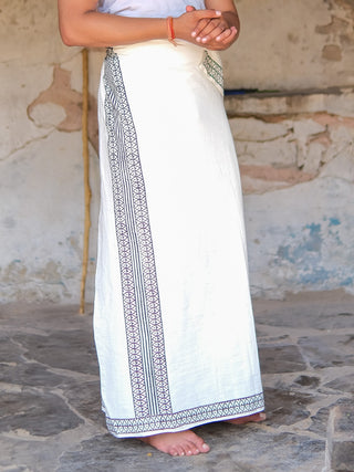 A person in a Nilaya Dhoti, crafted from premium cotton with a black block print design, stands barefoot on a stone floor before a rustic textured wall.