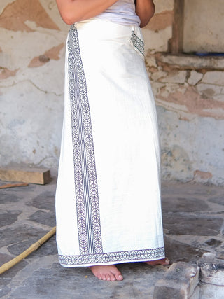 A barefoot person stands on a stone floor, hands clasped, wearing the Nilaya Dhoti—crafted from premium cotton with block print borders. The upper body is partially out of frame.