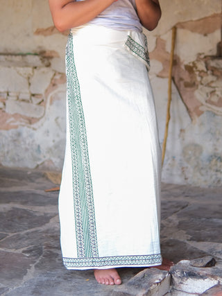 A person stands barefoot on stone flooring, dressed in the Nilaya Dhoti featuring premium cotton and green geometric block print borders, paired with a white top, set against a weathered textured wall.