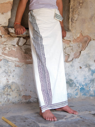 Wearing a Nilaya Dhoti made from premium cotton with a striking black border, a person stands barefoot beside an aged stone wall.