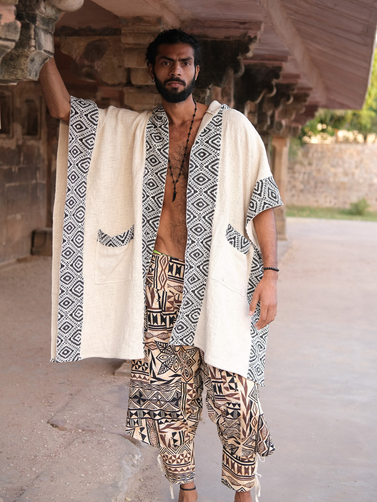 A man wears the Neer Poncho, a cream-colored block print garment with black and white geometric patterned borders and matching pockets, paired with patterned drop-crotch trousers.